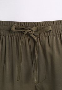 Ceinture élastique avec cordon noué sur un pantalon en tissu vert olive.