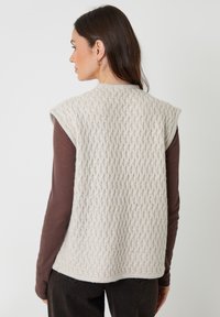 Stickad utanärmad väst i ljusbeige med ett texturerat mönster. Har en rund halsringning och mjukt tyg, bärs över en brun långärmad skjorta.