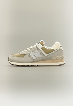 Beige en lichtgrijze New Balance sneaker met wit "N"-logo, witte veters en een gedempte zool tegen een effen achtergrond.