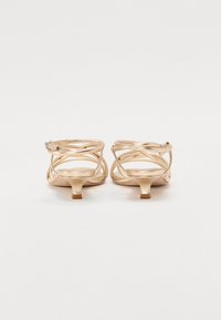 Chaussures à talons kitten dorées avec des lanières, présentant une silhouette élancée, des sangles croisées et un petit talon carré. Fini métallique lisse.