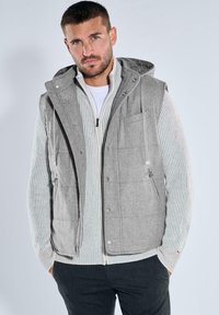 Mann trägt eine graue ärmellose Kapuzenweste über einem gestreiften Reißverschluss-Pullover und dunklen Hosen, steht mit den Händen in den Taschen vor hellem Hintergrund.