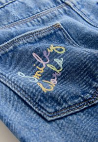 Kapsa z denimu s vícebarevným vyšívaným nápisem "Smiley World." Modrá barva s žlutými, růžovými a světle modrými akcenty.