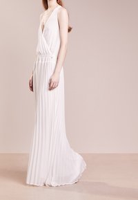 Kobieta w bezrękawnikowej, białej, plisowanej sukni maxi z głębokim dekoltem w kształcie litery V, stojąca na jednolitym, beżowym tle.