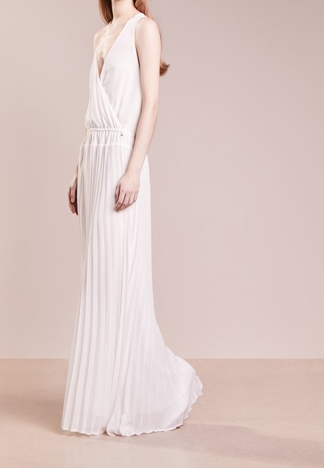Kobieta w bezrękawnikowej, białej, plisowanej sukni maxi z głębokim dekoltem w kształcie litery V, stojąca na jednolitym, beżowym tle.