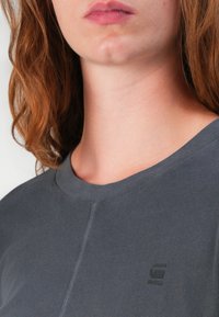 Grå t-shirt med rund hals och korta ärmar, tillverkad av mjukt tyg. Har en liten svart logotyp på vänster bröst och subtila sömdetaljer.