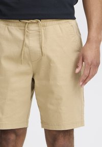 Beežid puuvillased lühikesed püksid, millel on elastne vöökoht paelaga, küljetaskud ja sirge lõige, nähtav vöökoht kuni reite keskosa.