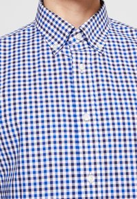 Camicia a quadri vichy bianca, blu scuro e blu chiaro; colletto button-down; bottoni bianchi; tessuto morbido con una consistenza strutturata.