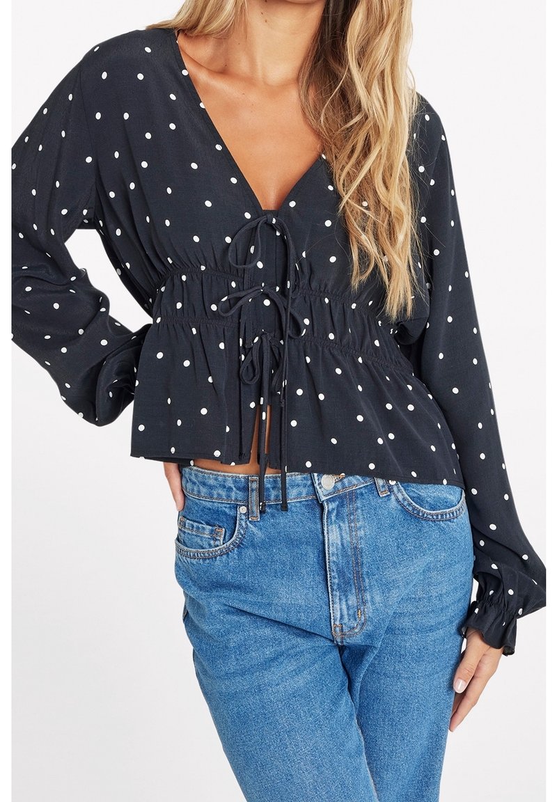 Sort bluse med hvide prikker, dyb V-udskæring, samlet talje og lange flæseærmer. Kombineret med lyseblå denimjeans.