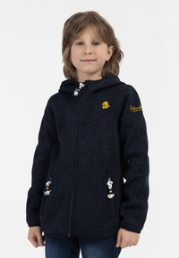Bambino che indossa una felpa con zip blu navy con un logo giallo e testo ricamato, in piedi su uno sfondo bianco semplice.