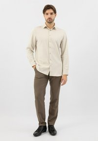Camicia button-up in tessuto beige chiaro, con maniche lunghe e colletto a punta, abbinata a pantaloni marroni e scarpe nere.