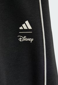 Crni sportski materijal sa žljebastom teksturom koji prikazuje bijele logotipe Adidas i Disney, naglašen uskim bijelim prugom sa strane.