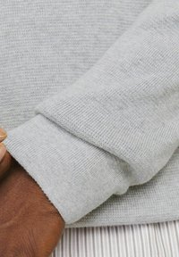 Polsino di un maglione lavorato a maglia grigio chiaro con design piegato, caratterizzato da una texture liscia e orlo a costine. Una mano poggia sul polsino.