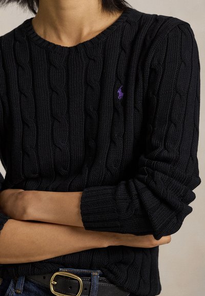 Henkilö, jolla on päällään musta kaapelineuletakki, jossa on violetti kirjailtu logo, kädet ristissä, yhdistettynä tummiin farkkuihin ja mustaan vyöhön, jossa on kullanvärinen solki.