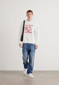 Felpa bianca con grafica rossa, jeans blu larghi e sneaker bianche. Il modello porta una borsa a tracolla nera e si trova su uno sfondo neutro.