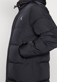 Zwarte pufferjas met capuchon, voorzien van een zilveren logo op de borst, ritszakken en een geborduurde textuur voor isolatie.