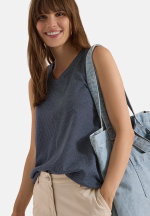 Femme souriante aux cheveux bruns porte un haut sans manches rayé bleu marine et un pantalon beige, tenant un grand sac bleu clair sur son épaule.