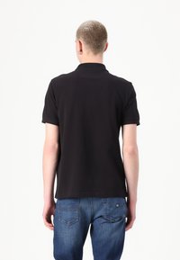 Tricou polo negru cu guler clasic și mâneci scurte. Fabricat dintr-un material neted, prezentat din spate, asortat cu blugi albaștri.