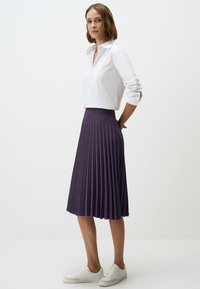 Gonna plissettata viola realizzata in tessuto leggero, abbinata a una camicia bianca con colletto. La modella è in piedi con le mani sui fianchi, indossando sneaker bianche.