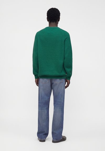 Homme debout de dos, portant un pull vert, un jean bleu et des chaussures noires sur un fond blanc uni.