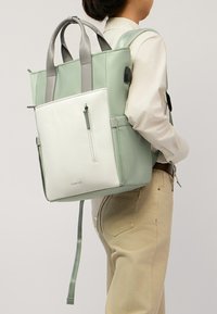 Zaino verde chiaro e bianco con finitura testurizzata, doppie maniglie superiori, spallacci regolabili e una tasca anteriore con zip.