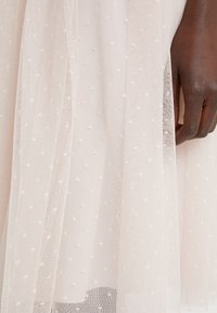 Tela transparente de color rosa claro con un patrón texturizado de pequeñas cruces blancas, caída suavemente, revelando una mano que sostiene la tela en el borde.