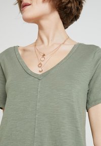 Zielona koszulka typu V-neck z miękkiego, teksturowanego materiału. Posiada krótkie rękawy i widoczne szwy wzdłuż środka. Warstwowe naszyjniki w złotym odcieniu podkreślają dekolt.