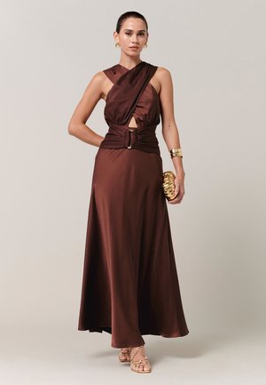 Vrouw draagt een mouwloze bruine satijnen jurk met overslag lijfje en brede riem, houdend een gouden clutch, gouden oorbellen, armband en sandalen.