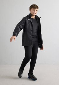 Dreng smiler iført sort sportsjakke, sorte leggings og fodboldstøvler, stående på ét ben mod en ensfarvet hvid væg.