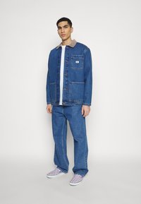 Blauwe spijkerjack met een beige corduroy kraag, een voorzak en gouden knopen, gecombineerd met blauwe spijkerbroek en paarse geruite instappers.