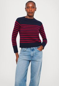 Meriharmaalla raidoitettu neulepusero, jossa on punaiset vaakaraitat, 3/4-pituiset hihat, ribbetyt hihansuut ja helma, yhdistettynä vaaleansinihin leveälahkeisiin farkkuihin.