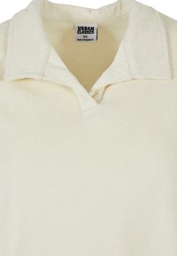 Cremefarget polo skjorte med teksturert stoff, krage som brettes over, og sydd placket. Merket sier "Urban Classics," størrelse XS.