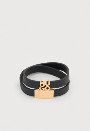 Svart läderarmband med två överlappande band, ett guldfärgat spänne och bokstäverna "HUGO" tydligt visade i metallisk finish.