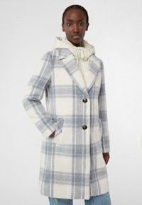 Cappotto a quadri in misto lana color crema e grigio chiaro, con colletto, due tasche frontali e due bottoni marroni. Indossato sopra un hoodie chiaro.