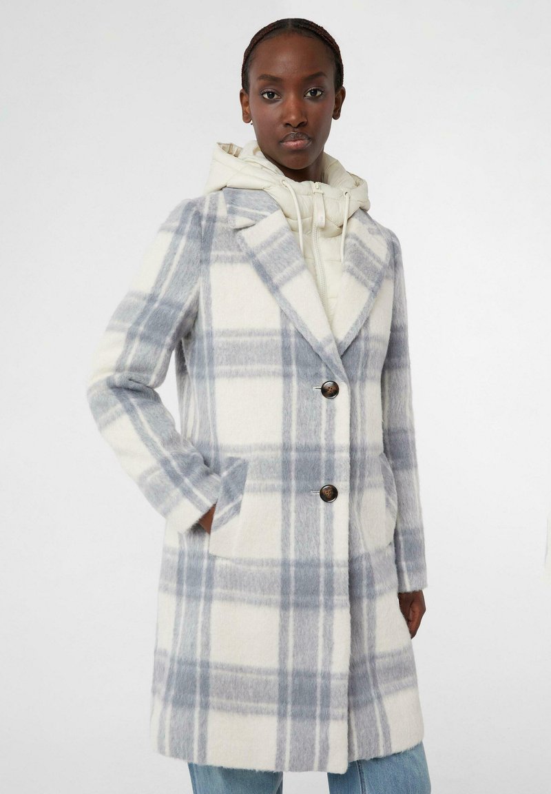 Cappotto a quadri in misto lana color crema e grigio chiaro, con colletto, due tasche frontali e due bottoni marroni. Indossato sopra un hoodie chiaro.