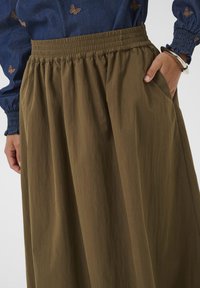 Kvindelig iført en brun skørtoverdel med samlet talje og en blå langærmet bluse med broderede sommerfugle, hånd i skørtekanten, sølv armring.