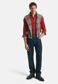 Chemise rouge à boutons, pull en tricot gris drapé sur les épaules, jean bleu marine et chaussures noires. Tissu finement texturé et couleurs contrastées.