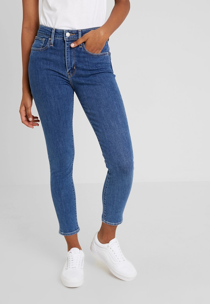 Personne portant un jean slim bleu et des baskets blanches, debout avec une main dans la poche devant un fond uni gris clair.