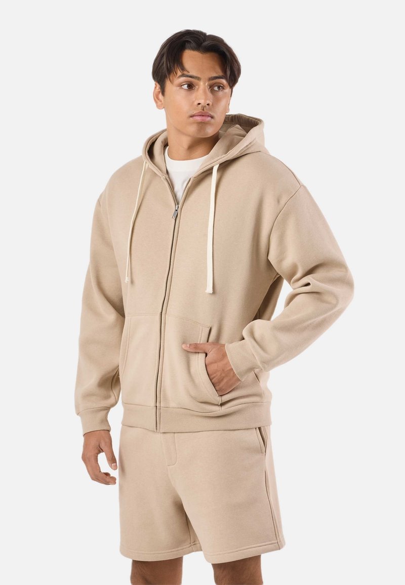 Jeune homme portant un sweat à capuche beige zippé avec cordons et un short assorti, debout avec une main dans la poche, regardant sur le côté.