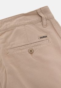 Pantaloni in cotone beige con una texture liscia, dotati di una tasca posteriore, chiusura con bottone e un'etichetta con logo "GUESS" nera.