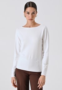 Maglione bianco a maniche lunghe con scollo a barca e trama a coste, con bottoni decorativi sulle maniche e un discreto logo sul fondo.