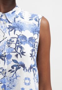 Blusa a maniche corte bianca con motivo floreale blu, caratterizzata da un'apertura con bottoni e colletto classico. Tessuto dalla texture liscia.