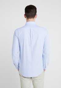 Niebiesko-biała koszula w paski z długim rękawem, kołnierzykiem na guziki, zaszewką z tyłu i zaokrąglonym dołem. Materiał wydaje się gładki, lekki i przewiewny.