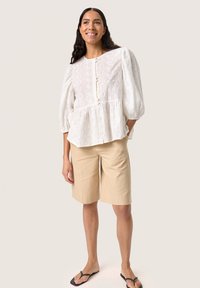 Blusa bianca testurizzata con maniche a sbuffo e chiusura a bottoni, abbinata a pantaloni corti beige fino al ginocchio e infradito nere.