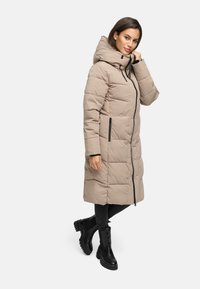 Beige gewatteerde lange jas met hoge kraag, rits aan de voorkant en zijzakken; gecombineerd met zwarte jeans en enkelboots. Gladde textuur.
