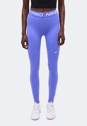 Γυναίκα με μοβ κολάν Nike Pro και λευκά αθλητικά παπούτσια, στέκεται μπροστά σε ένα απλό φωτεινό φόντο.