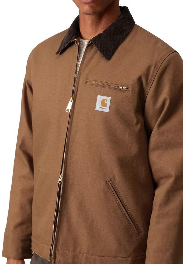 Brown zip-up jacket with dark corduroy collar, zip chest pocket, and Carhartt logo patch on left side worn by a person.