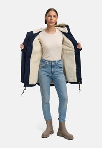 Navy jacket with a fuzzy cream lining, worn open. The person is in light blue jeans and beige ankle boots. Simple design, practical materials.