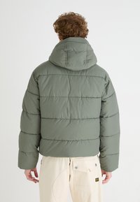 Puffer jacket in olive green with horizontal stitched design and a large hood. The fabric appears soft and insulated for warmth.