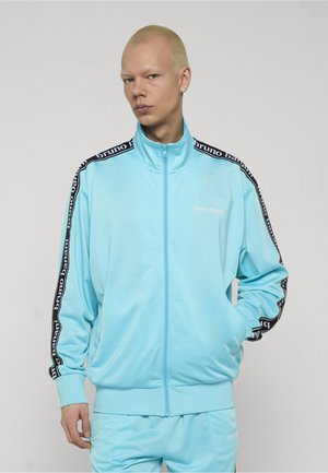 Person med kort blondt hår iført en lyseblå Bruno Banani træningsdragt med sorte logostriber på ærmerne og hænderne i lommerne.