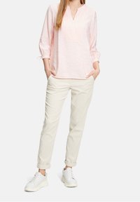 Haut rose clair avec un col en V et des plis à l'avant, associé à un pantalon beige retroussé et des baskets blanches. Tissu texturé, design décontracté.